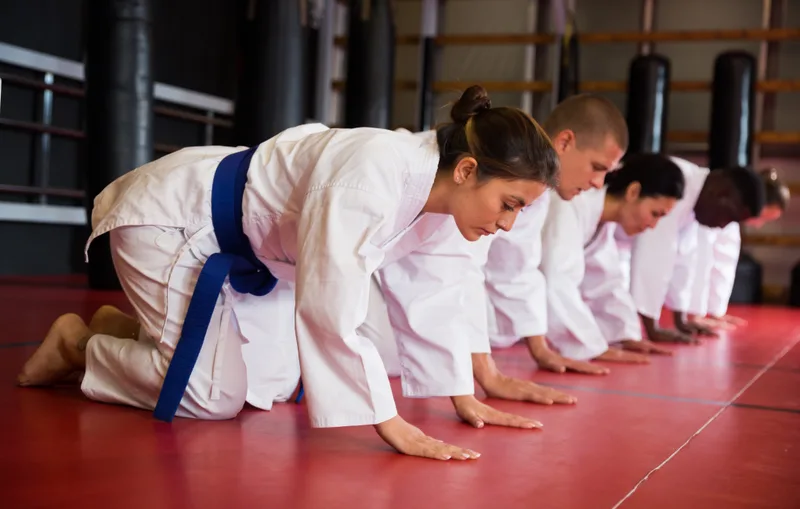 Row of adults in white gis kneeling in a quiet, focused line on a red mat at the start of class
