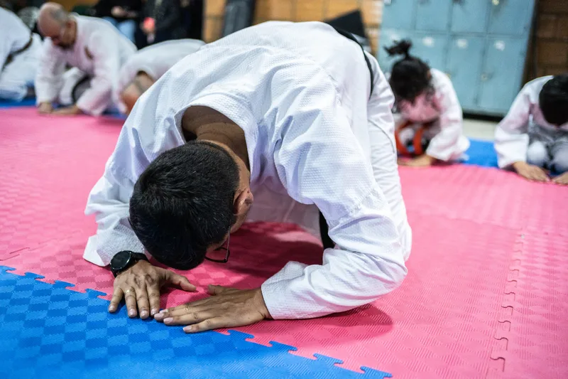 Student in a white gi bowing deeply on interlocking mats while younger classmates kneel in the background