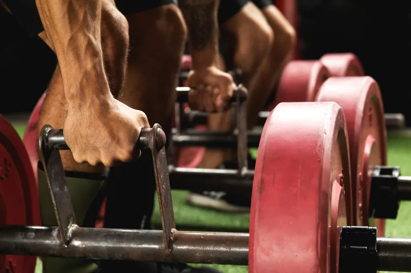 Close-up of two athletes' hands gripping farmers-carry handles loaded with red plates on green turf