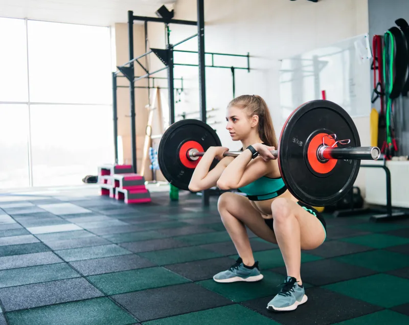 Member holding a loaded barbell in the front-rack position at the bottom of a squat, in a bright gym with a rig behind her