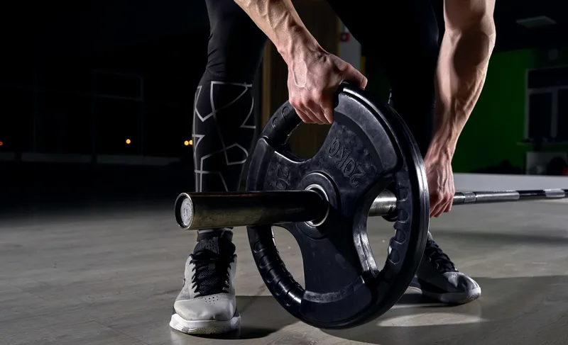 Lifter crouching to load a black iron plate onto a barbell on a dark gym floor