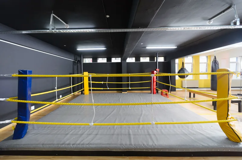 Boxing ring with yellow ropes inside a gym studio, heavy bags and a speed bag hanging along the far wall