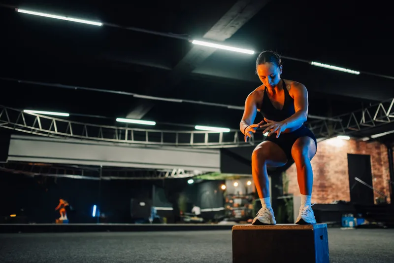 Athlete checking her watch while crouched on a plyo box in a dark warehouse gym strung with strip lights