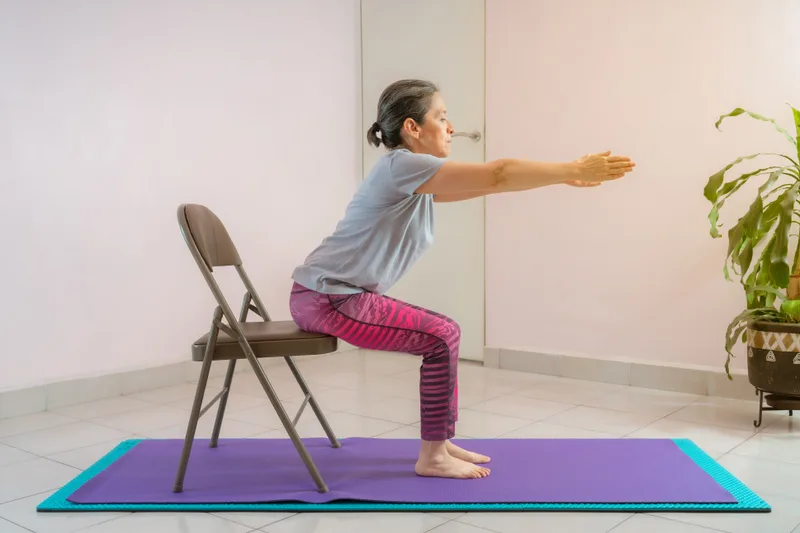An older student practises a supported chair pose on a mat with a folding chair behind her
