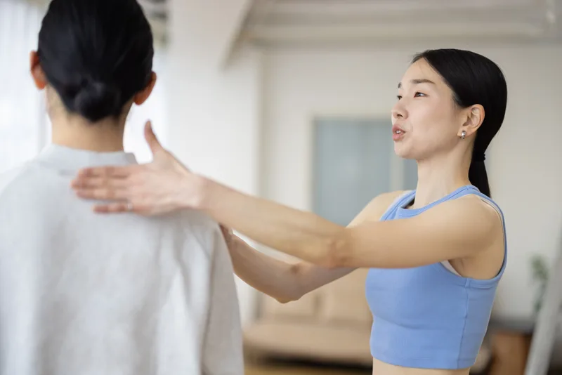 A teacher gently checks a student's shoulder position while explaining the pose