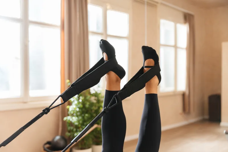 Student's legs in dark grip socks pressing overhead into reformer straps in a bright studio