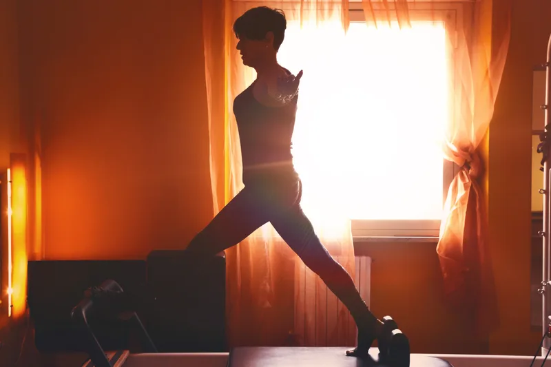 Silhouette of a student holding a deep lunge on a reformer against a glowing curtained studio window
