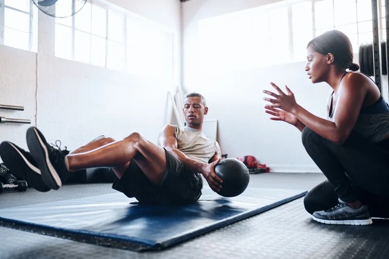 A coach kneeling beside a client on a gym mat, coaching him through medicine-ball core work in a sunlit gym