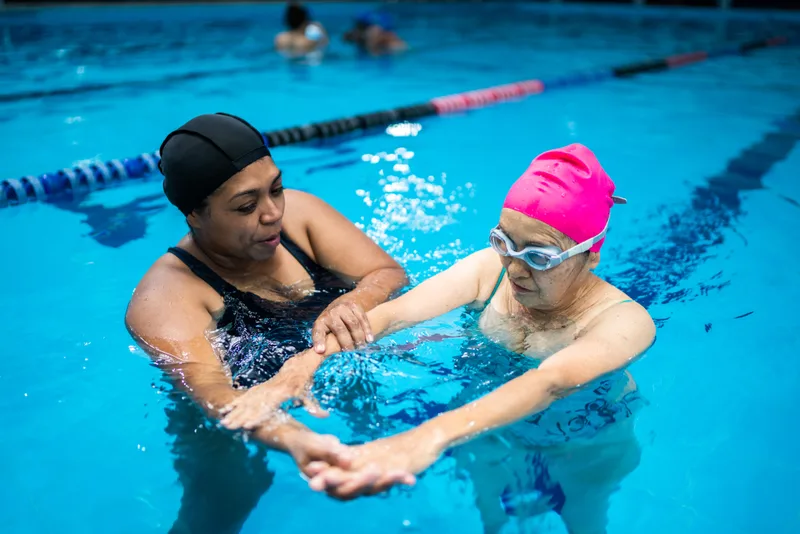 Instructor in the pool gently supporting an adult student's arms as she practises reaching forward in the water