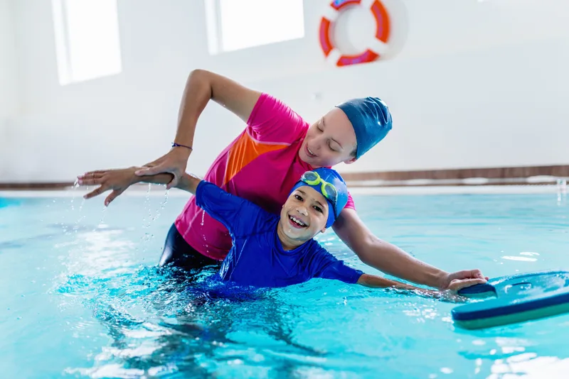 Instructor supporting a smiling boy as he reaches for a kickboard during a one-on-one swim lesson