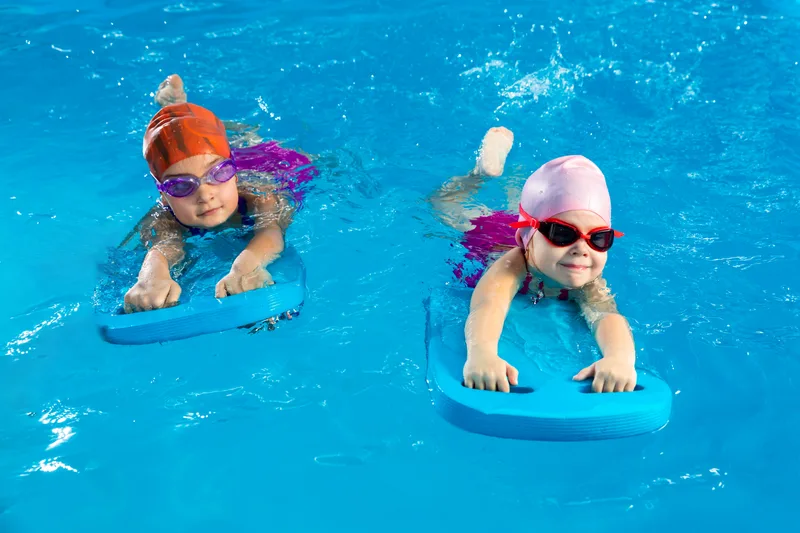 Two young children in swim caps and goggles kicking across the pool on blue foam kickboards