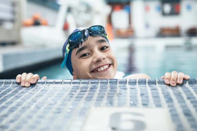 Boy in a swim cap with goggles pushed up grinning at the pool edge after a practice swim
