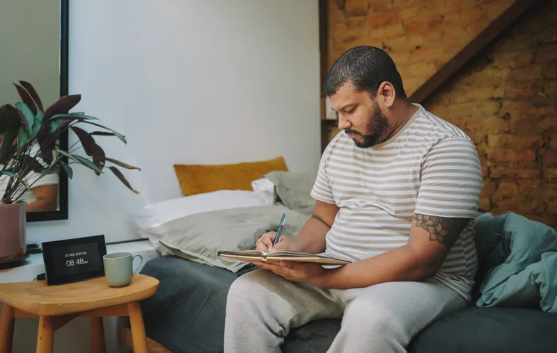 A man sitting on a bed at home writing in a notebook