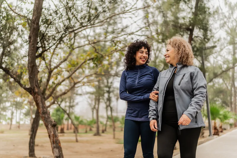 A mother and her adult daughter walking arm in arm and laughing in a park