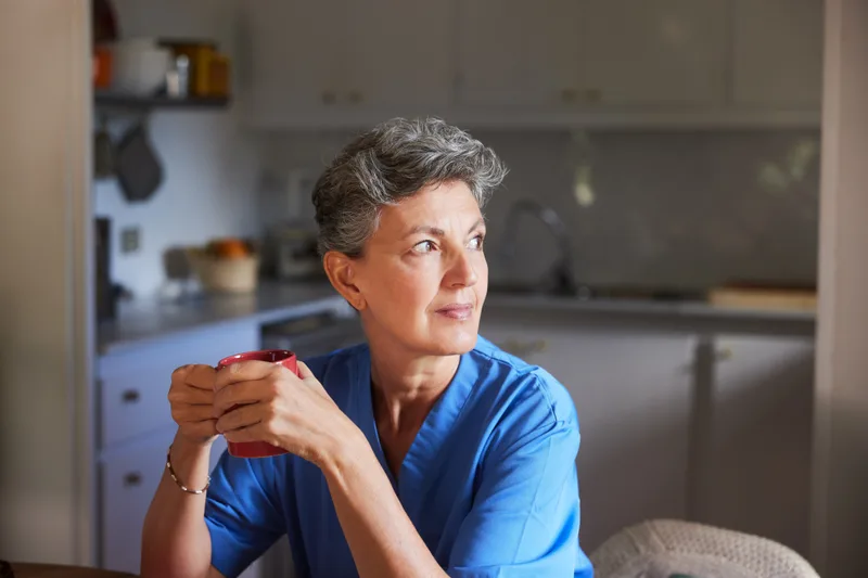 A nurse in blue scrubs holding a mug and looking out of a kitchen window