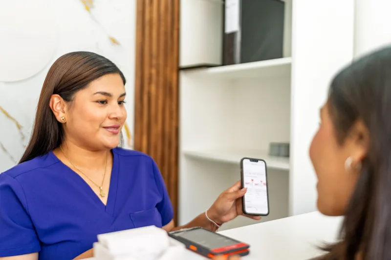 Treatment coordinator in blue scrubs showing a patient a payment plan on a phone at the front desk