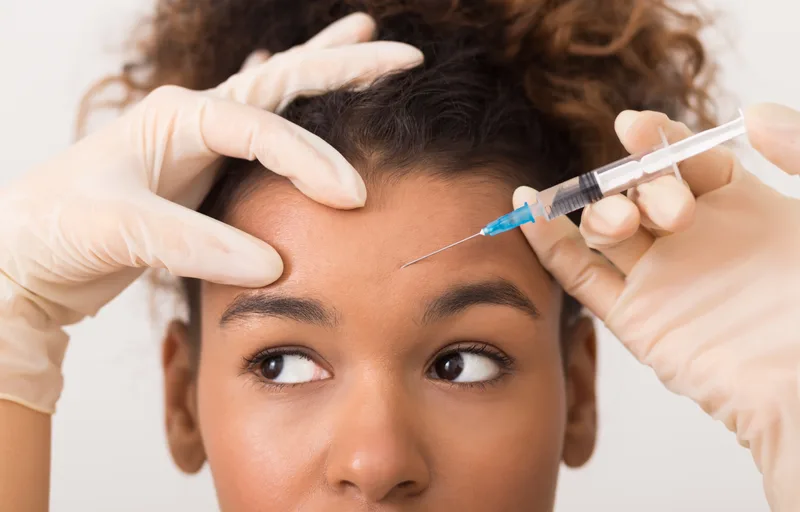 Gloved hands of a physician steadying a patient's brow while placing a botulinum toxin injection in the forehead