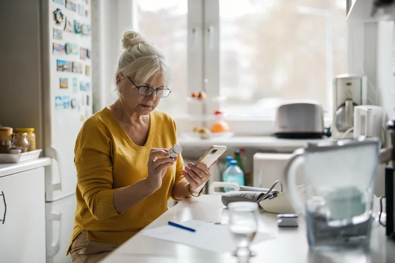 A member at her kitchen counter checking a medication bottle against her phone, home blood-pressure monitor at hand
