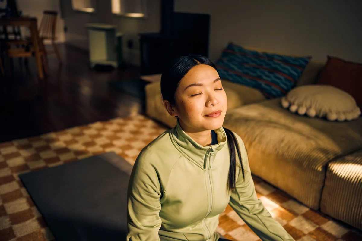 A woman sitting cross-legged in warm evening light at home, eyes closed, taking a quiet breath
