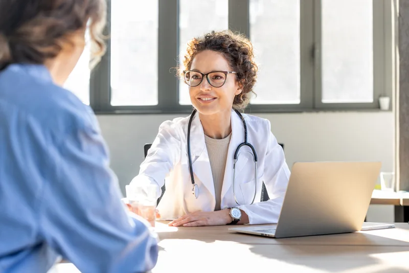 Physician in a white coat talking a patient through a plan across a desk in a bright consultation room