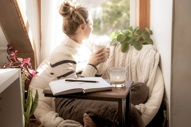 Woman journaling with a mug of tea in a cozy window nook beside a leafy plant