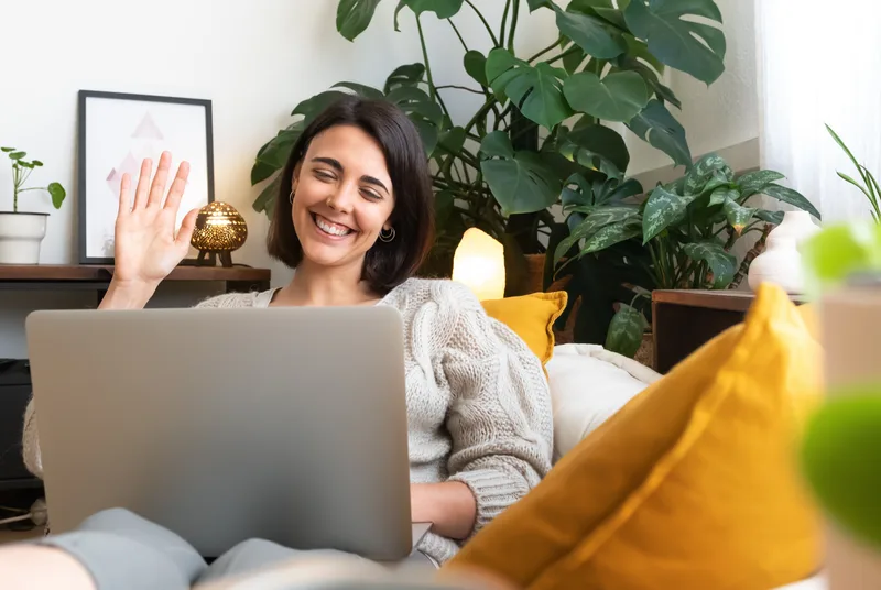 Woman waving hello to her therapist over a laptop video call from her plant-filled living room