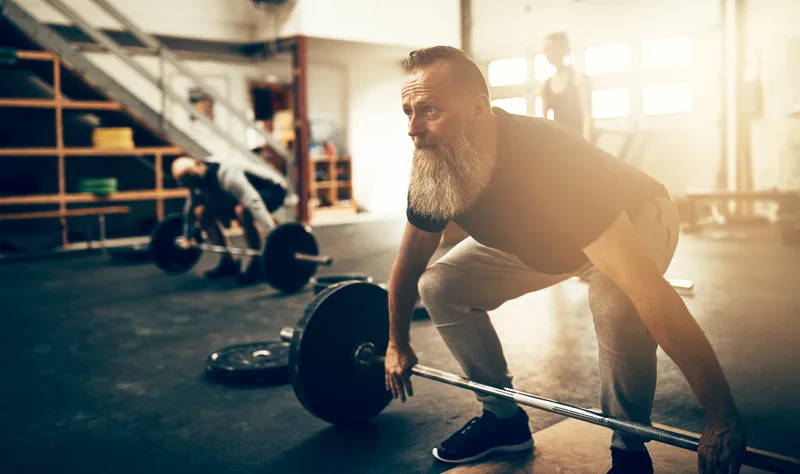 A bearded man in his fifties sets up for a barbell lift in a sunlit gym