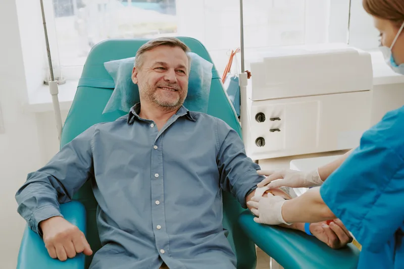 A middle-aged man in a blue shirt smiles in a reclining chair while a gloved technician draws blood from his arm