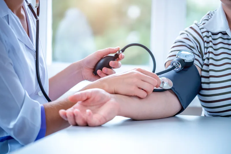 A clinician in a light blue coat takes the blood pressure of a man in a striped shirt using an arm cuff and stethoscope