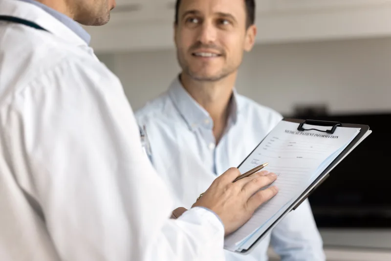 A clinician with a clipboard going over intake answers with a smiling male patient in a bright consult room