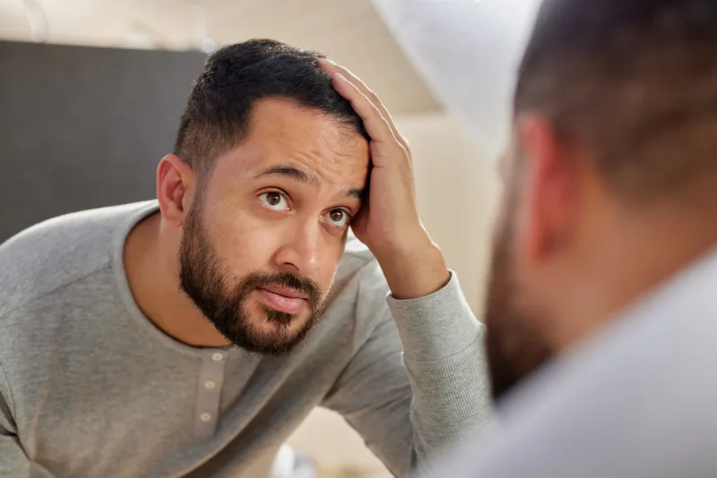 A bearded man runs a hand back through his hair while checking his hairline in a bathroom mirror