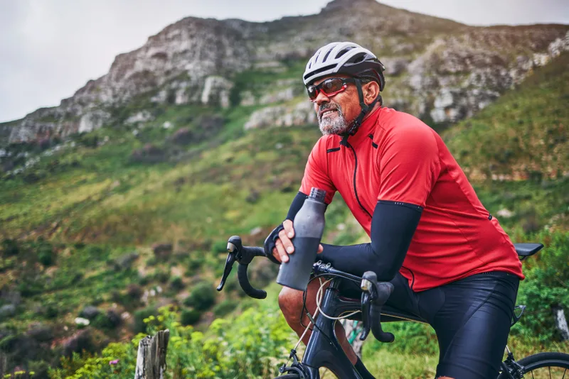 A bearded man in a red cycling jersey takes a drink on a road bike in green hills