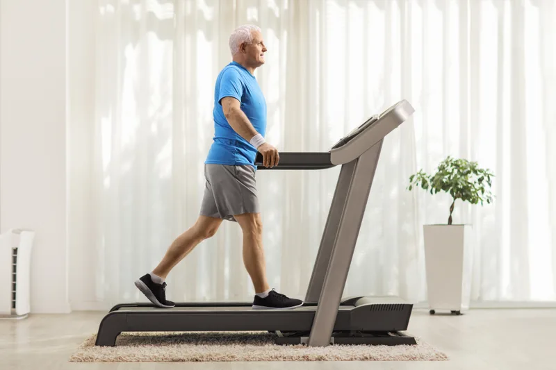 A grey-haired man in a blue t-shirt walking on a treadmill in a bright room with sheer curtains