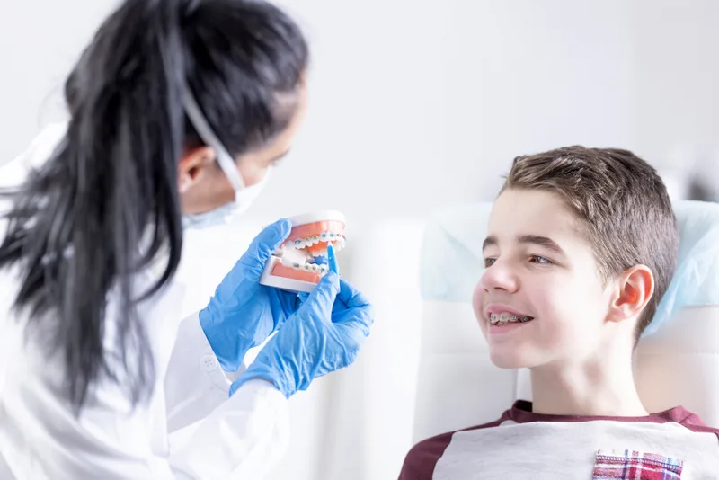 Orthodontist showing a young boy a teeth model with braces while he smiles in the chair