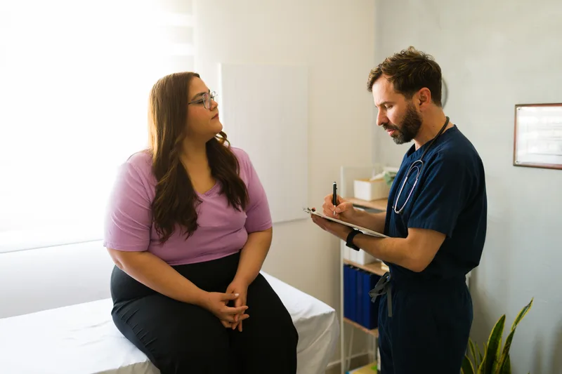 A patient sits on an exam table talking with a clinician who takes notes during a consultation