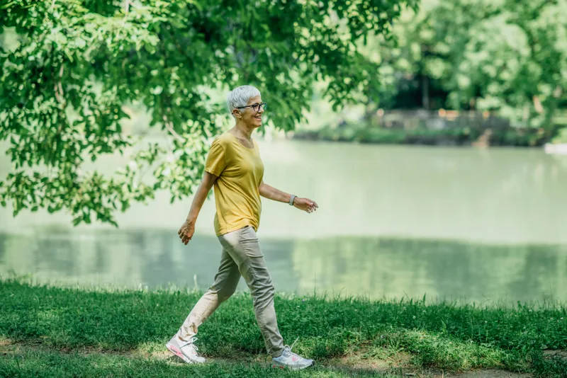 An older woman in a yellow shirt walks on the grass beside a lake