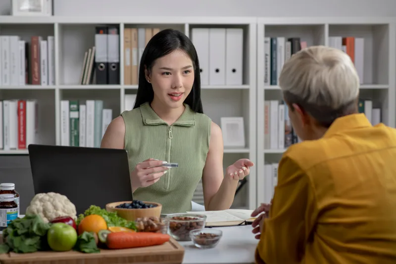 A dietitian talks through a meal plan with an older client at a desk with fresh vegetables
