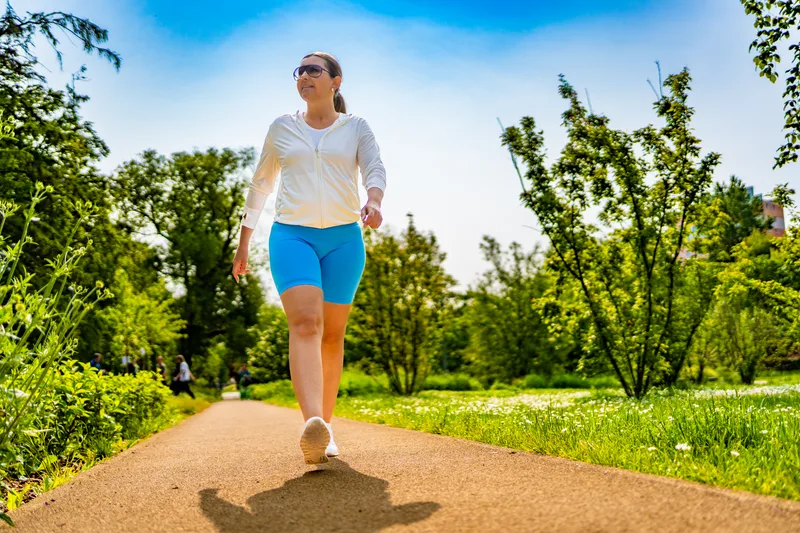 A woman walks along a sunny tree-lined path in a park