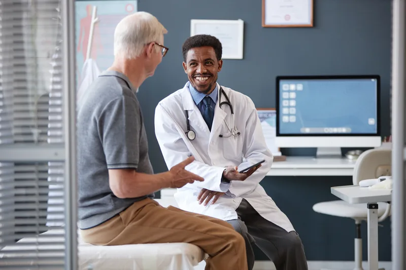 A smiling physician with a tablet talks with an older patient in a bright exam room