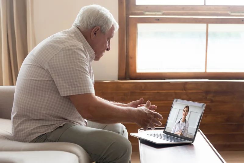 An older man at home talks with a doctor over a laptop video call at his coffee table