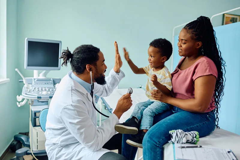 A doctor high-fives a delighted toddler sitting on his mother's lap in a mint-green exam room