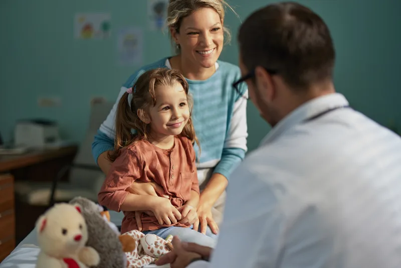 A young girl with pigtails sits calmly with her mother while a doctor kneels to talk with her, plush toys nearby