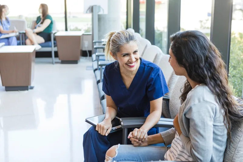 A nurse in navy scrubs chatting with a seated patient in the bright, glass-walled Northland lobby