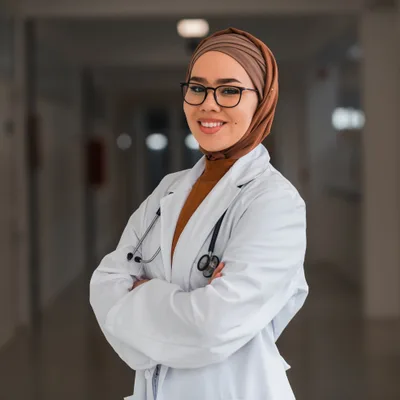 Dr. Amina Suleiman, a physician wearing a headscarf, glasses, and a white coat, standing in a clinic corridor