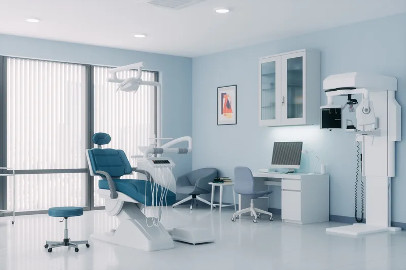 A bright consult room with a blue dental chair, a desk with a screen, and a panoramic scanner by the wall