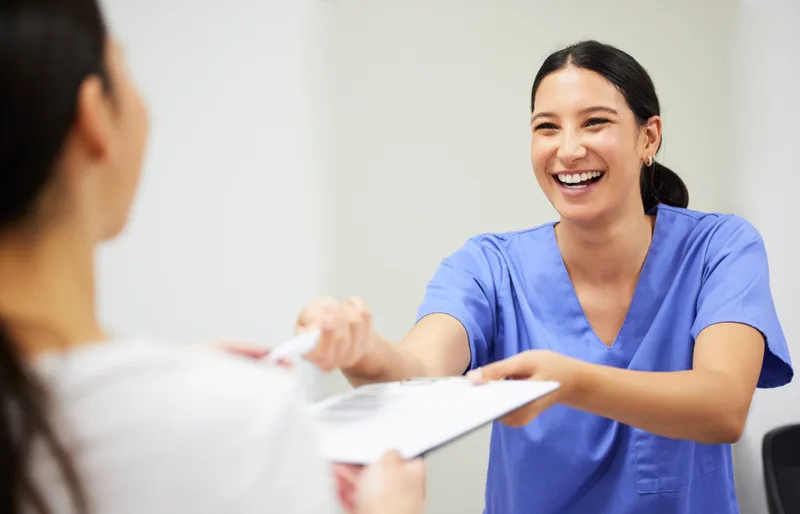 A smiling coordinator in blue scrubs hands paperwork to a patient across a desk