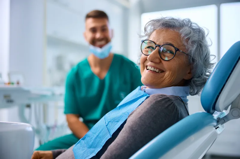 Smiling older woman in glasses relaxing in a dental chair while a clinician in green scrubs waits behind her