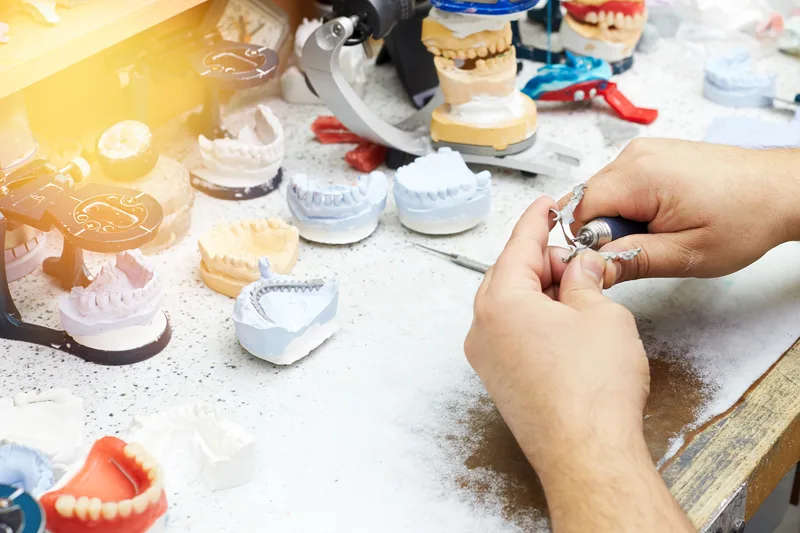 A dental lab technician's hands finishing a small restoration at a bench covered in tooth models and dentures