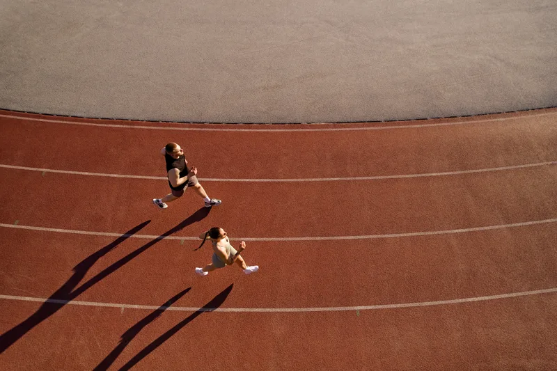 Two athletes running on a red track seen from above, with long evening shadows