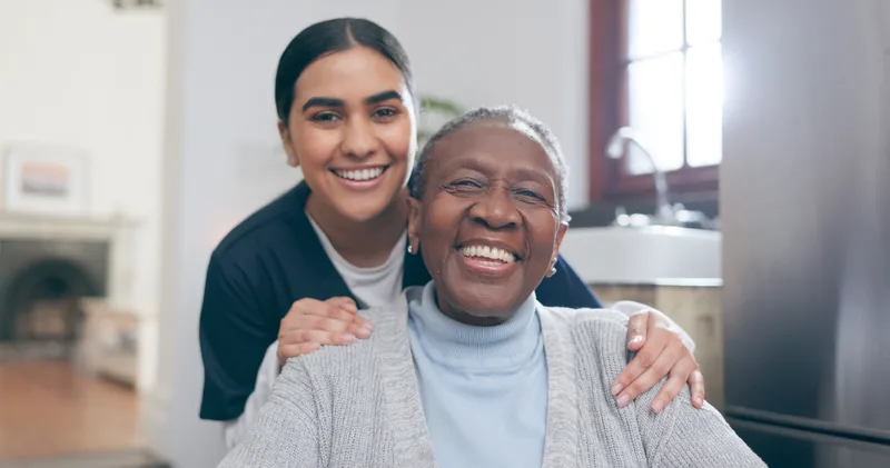 A smiling care partner stands behind a seated resident with her hands on her shoulders